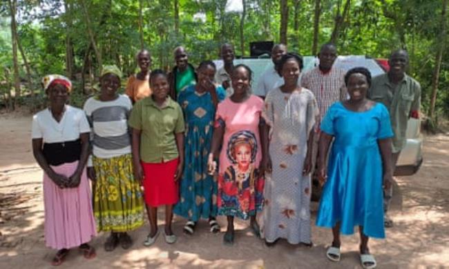 Two rows of people smile at the camera. They are standing in aclearing amo<em></em>ngst trees and behind them is a car covered with A3 paper bearing notes from a training session